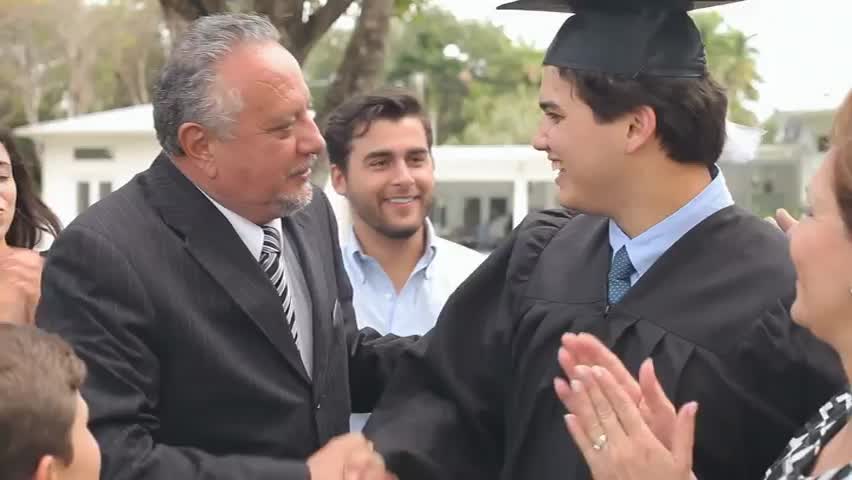 A graduate in cap and gown shaking hands with an older relative while the family claps
