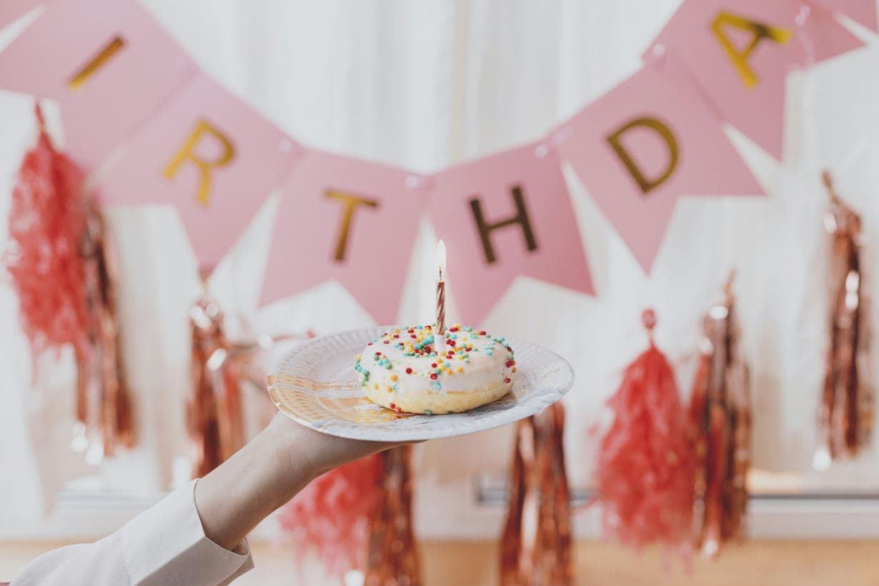 A doughnut with a lit birthday candle held up in front of a pink happy birthday banner